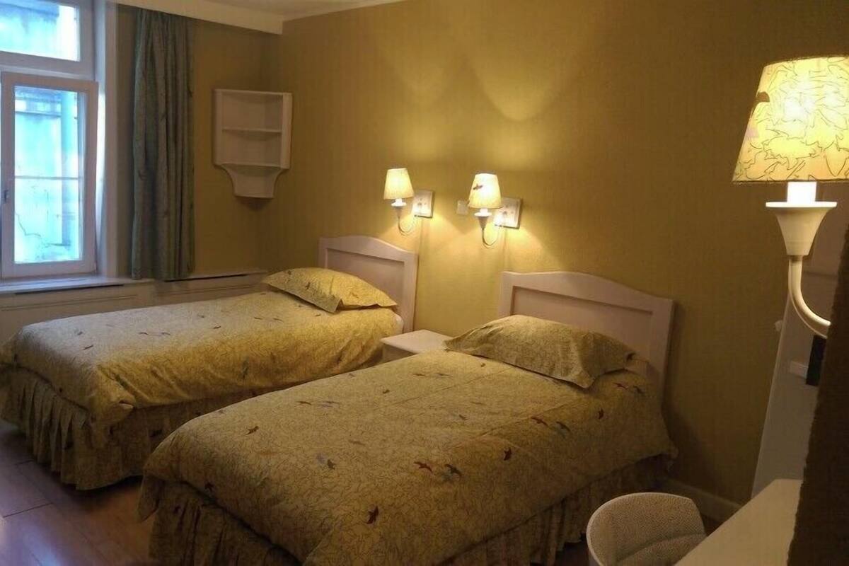 A hotel room with two single beds, a window with curtains, a wall-mounted shelf, and a bedside lamp.