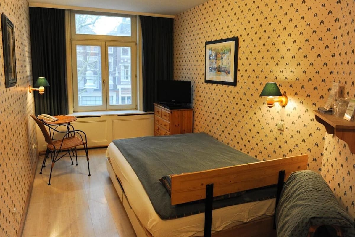 A bedroom with a bed, a desk with a chair, a television, and a window with curtains.