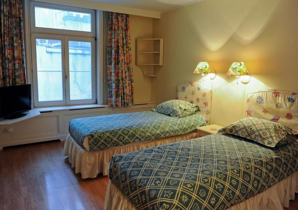 A room with two beds, a TV, a window with curtains, and two wall-mounted lamps.