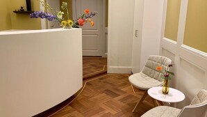 A reception area with a curved white counter, a small round table with a vase of flowers, and two upholstered chairs. - Imperial Hotel Rembrandtplein  (Amsterdam)