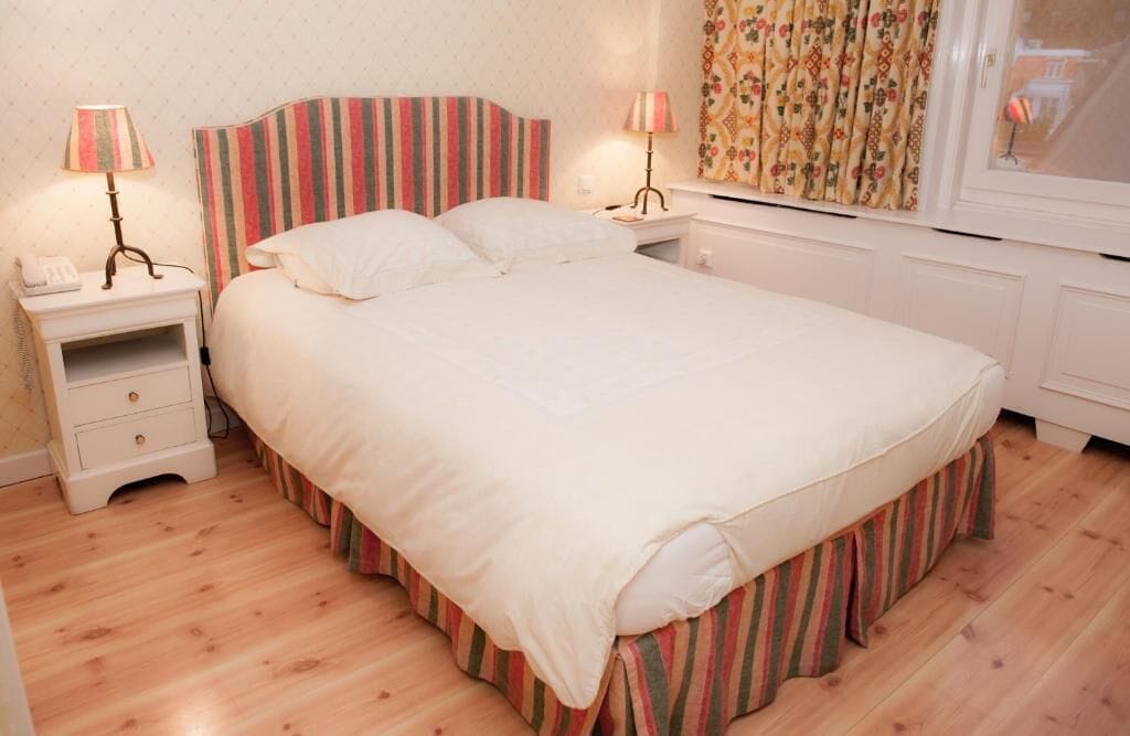 A neatly made bed with striped bedding, two bedside tables with lamps, and a window with patterned curtains.