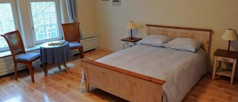 A bedroom with a wooden bed, two nightstands, a chair, a small table, and a window with a view of buildings.