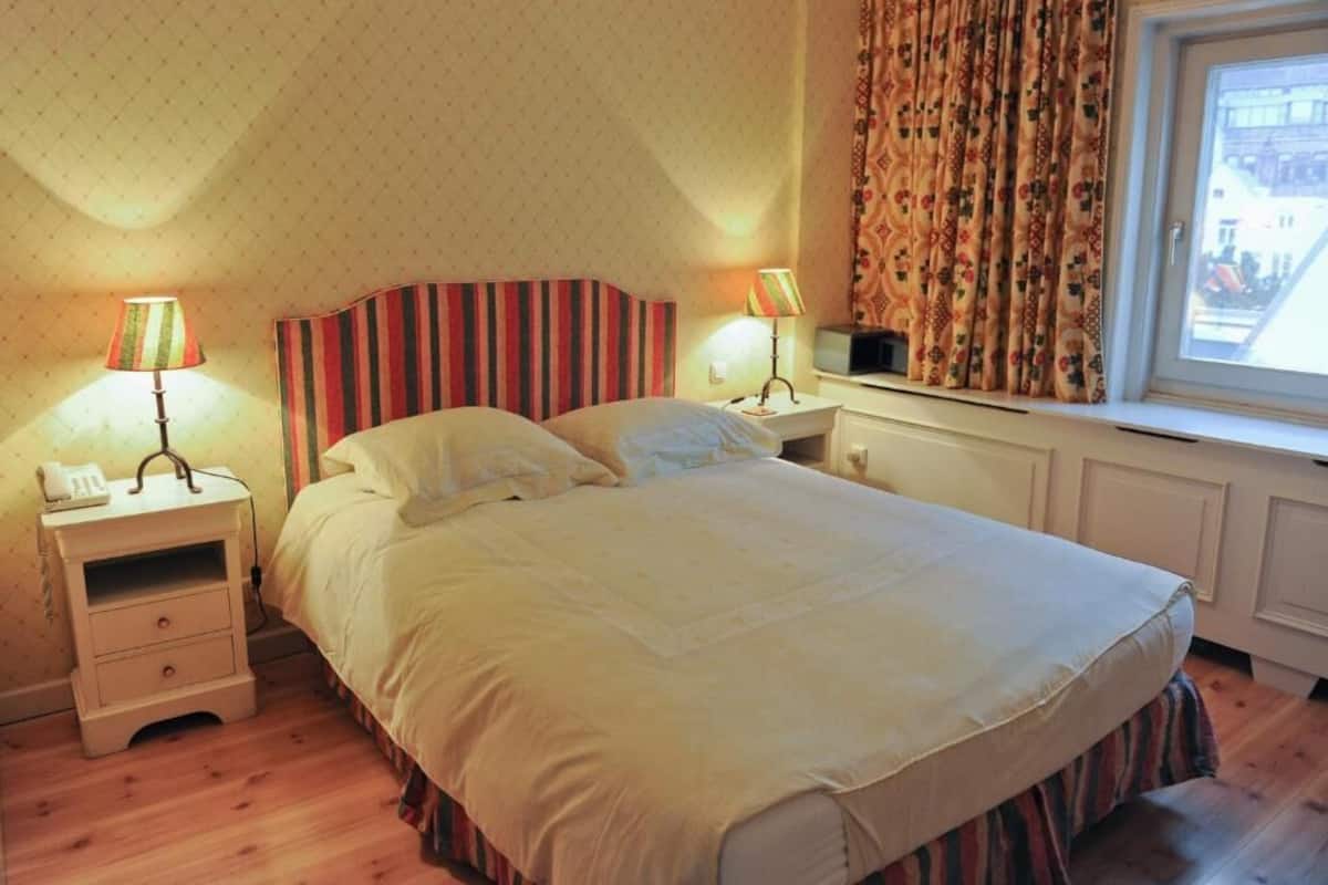 A bedroom with a bed, two bedside tables with lamps, a window with curtains, and a radiator.