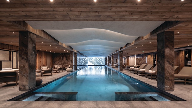 Une piscine intérieure moderne offrant une vue dégagée sur l’extérieur, avec des poutres en bois au plafond et des espaces de détente dotés de sièges.