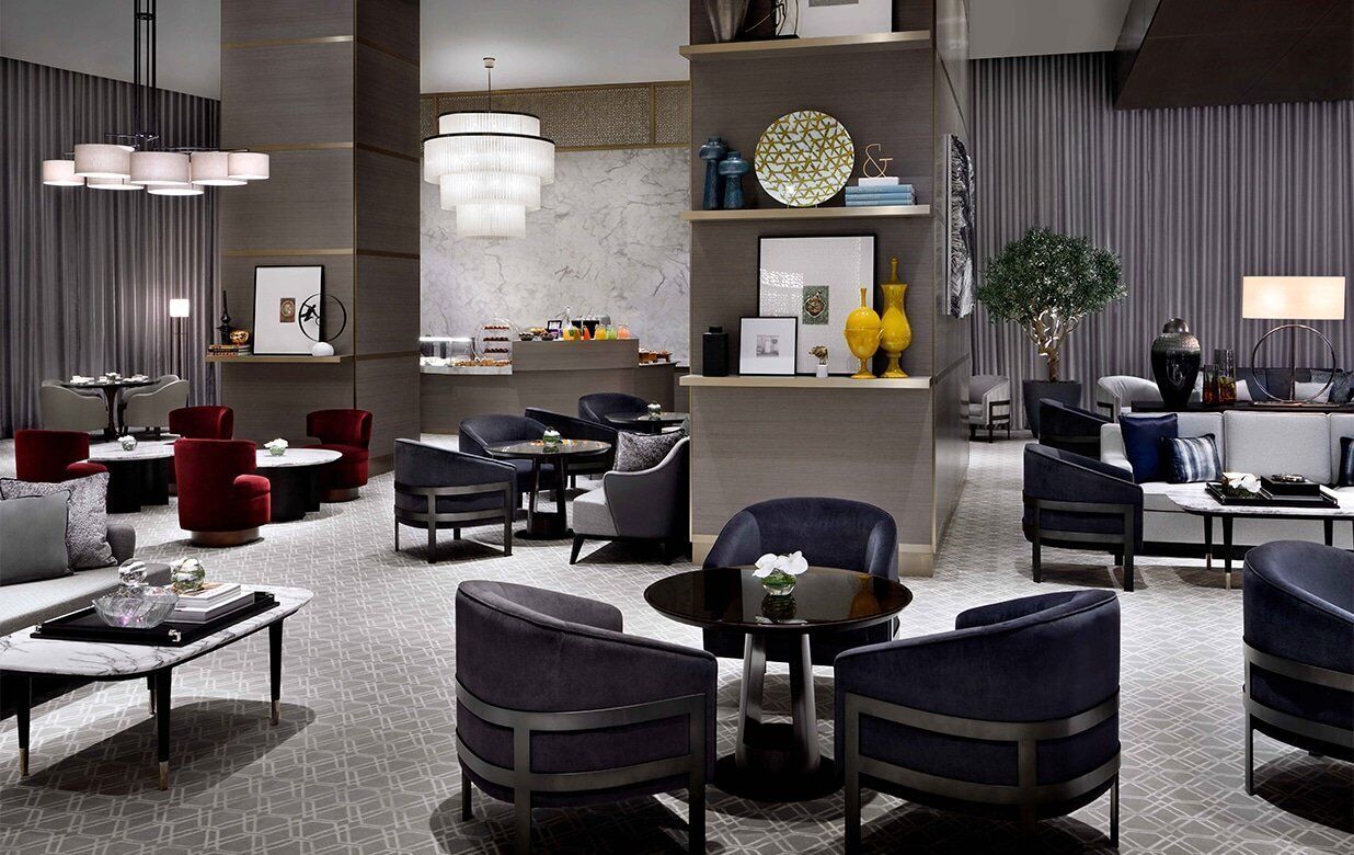 A modern hotel lobby with a mix of seating areas, including round tables and sofas, and a central bar area with a TV and bar stools.