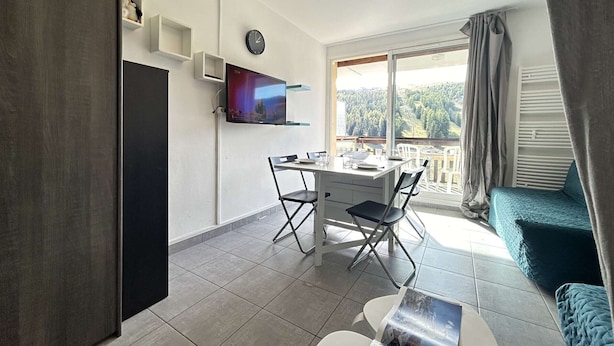 En modern lägenhet med ett matområde, en TV som är monterad på väggen, en balkong med utsikt över skogen och en mysig sittgrupp.