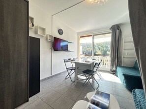 En moderne lejlighed med spiseplads, fjernsyn ophængt på væggen, en balkon med udsigt til skoven og et hyggeligt opholdsområde.