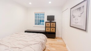 Un dormitorio con una cama, un televisor sobre un soporte negro, una ventana y un cuadro en la pared.