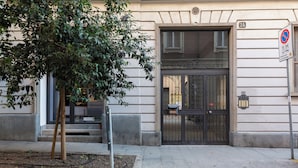 Une entrée d’immeuble avec un portail noir, un arbre au premier plan et un panneau indiquant qu’il est interdit de se garer.