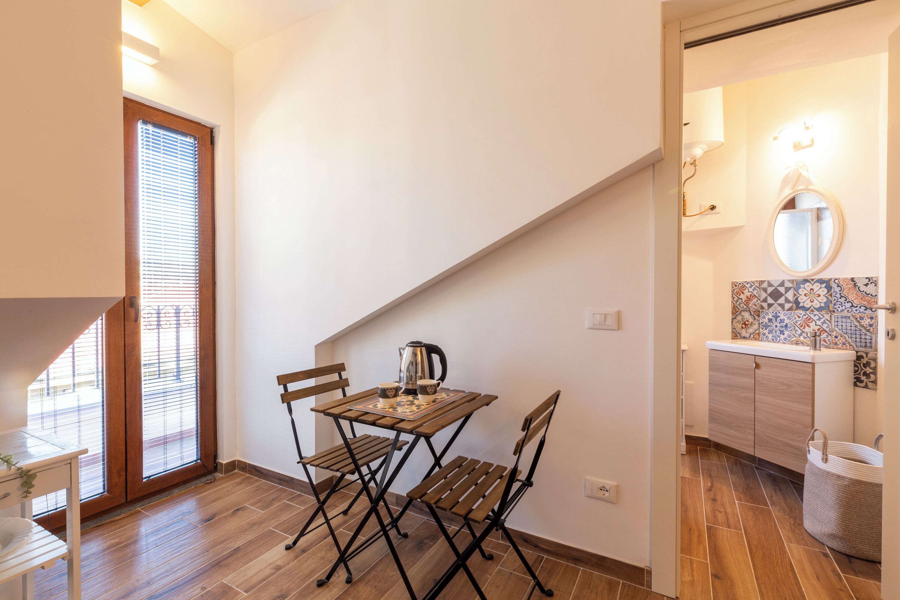 A small room with a wooden table and chairs, a kettle, and a window with blinds.