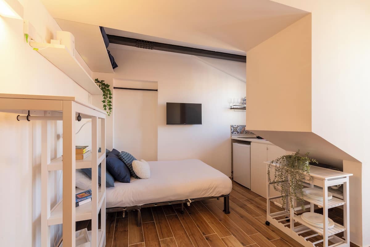 Un dormitorio pequeño con cama, un armario blanco, un escritorio con una planta y un televisor en la pared.