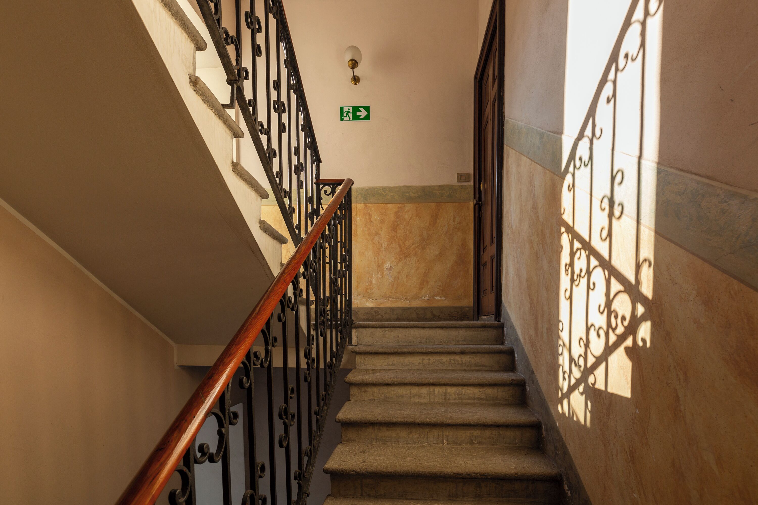 Una escalera con barandilla de madera y balaustradas de metal negro, que conduce a una plataforma con un letrero de salida de emergencia verde.