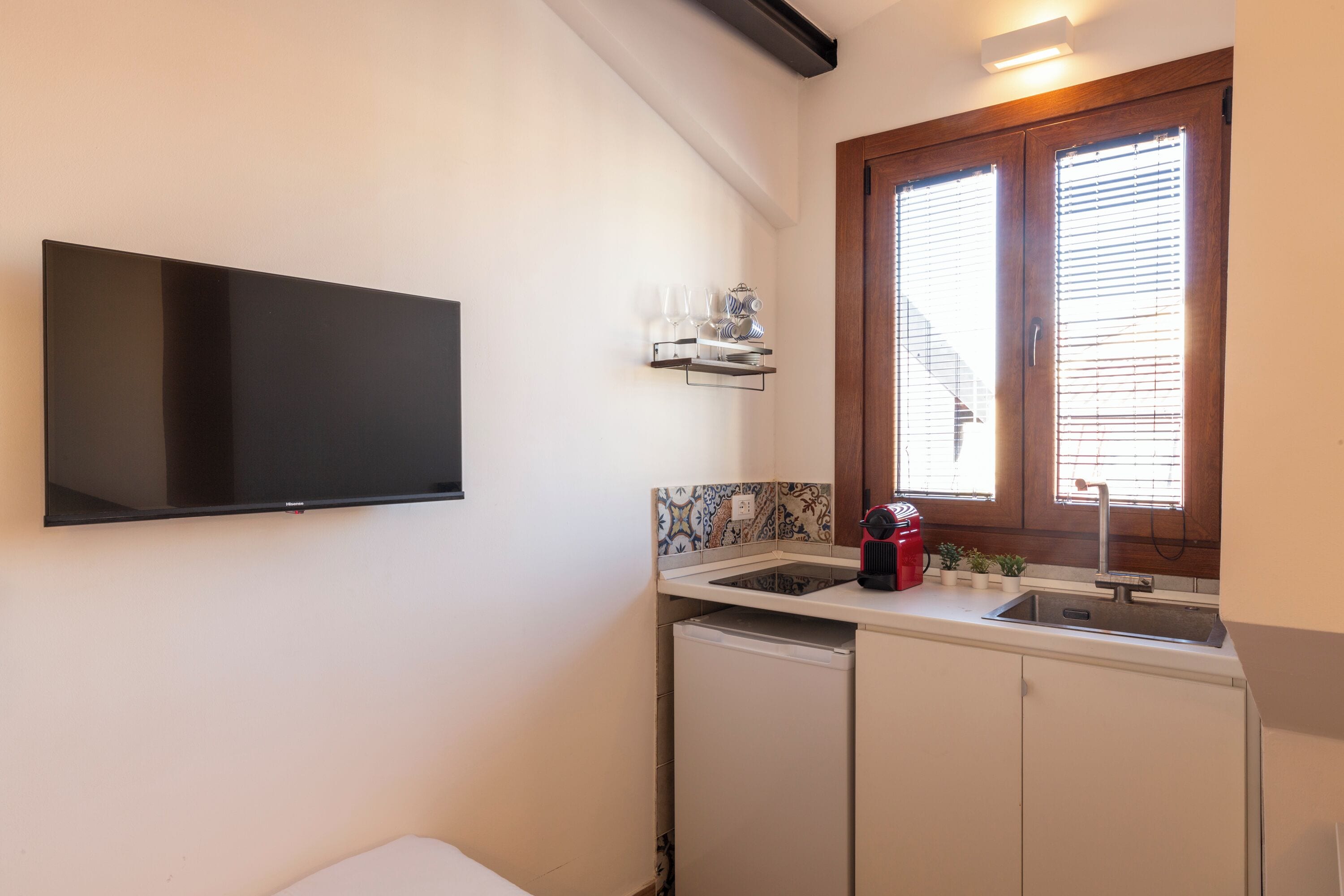 A compact kitchen with a built-in TV, a window with blinds, a sink, and a small countertop with a coffee maker.