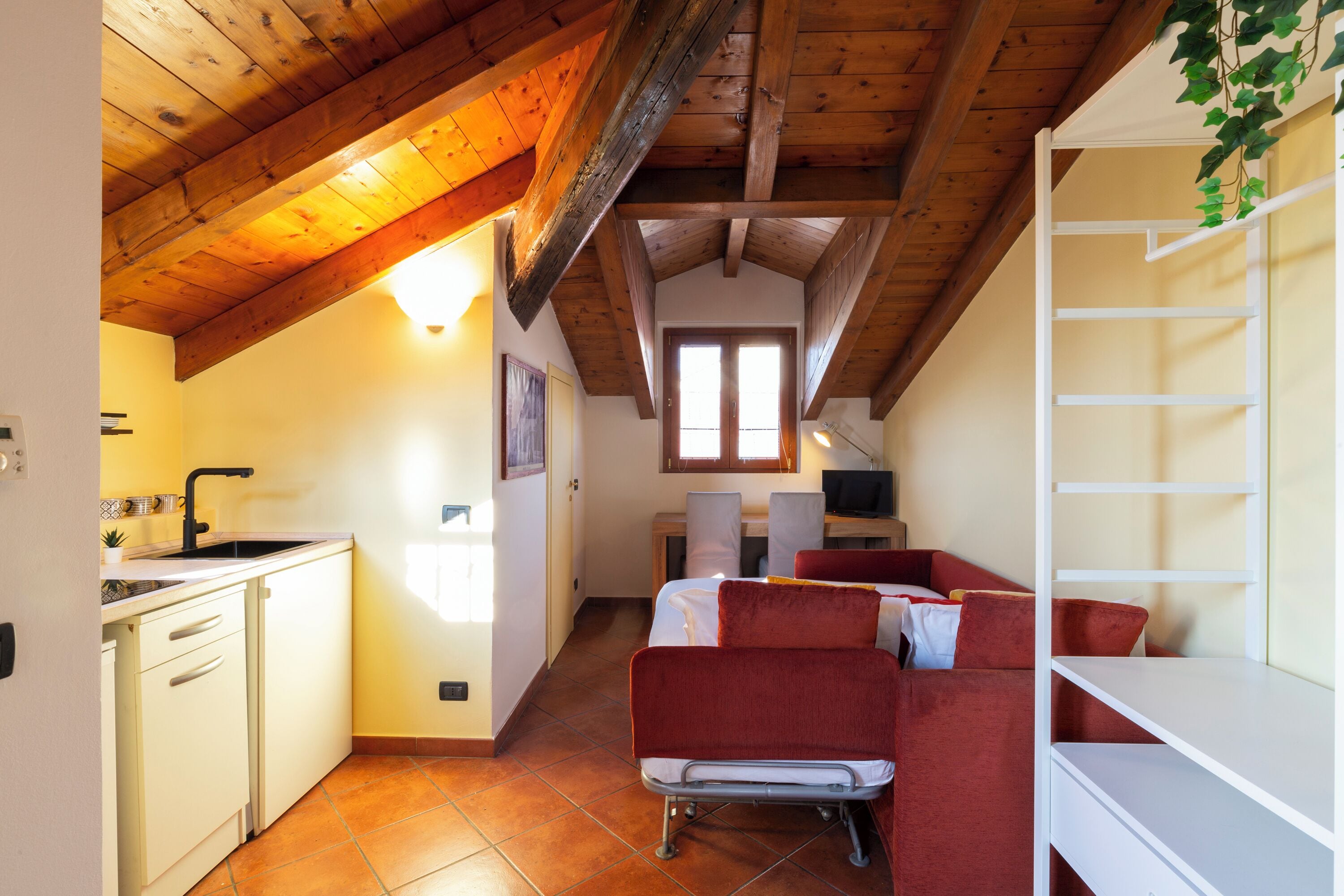 A compact kitchen with a sink and cabinets, a red sofa in the living area, and a white shelving unit.
