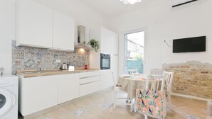 Eine moderne Küche mit weißen Schränken, einer gefliesten Spritzschutzwand und einer integrierten Waschmaschine. Es gibt einen Essbereich mit Tisch und Stühlen, sowie ein an der Wand befestigtes Fernsehgerät.