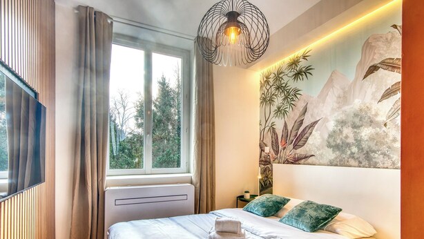 Une chambre moderne avec un grand lit, une fenêtre avec des rideaux, un luminaire fixé au mur et une fresque murale sur le thème de la nature.