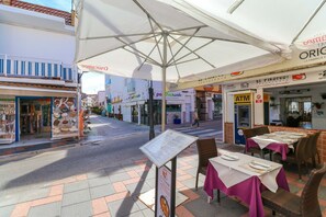 An outdoor café with tables and chairs, a menu stand, and various storefronts in the background. - La Cala beach apartment (La Cala de Mijas)