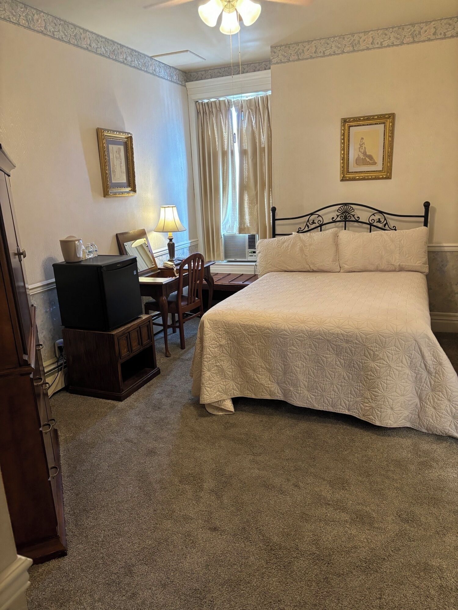 A hotel room with a bed, a desk with a lamp, a television, and two framed pictures on the wall.