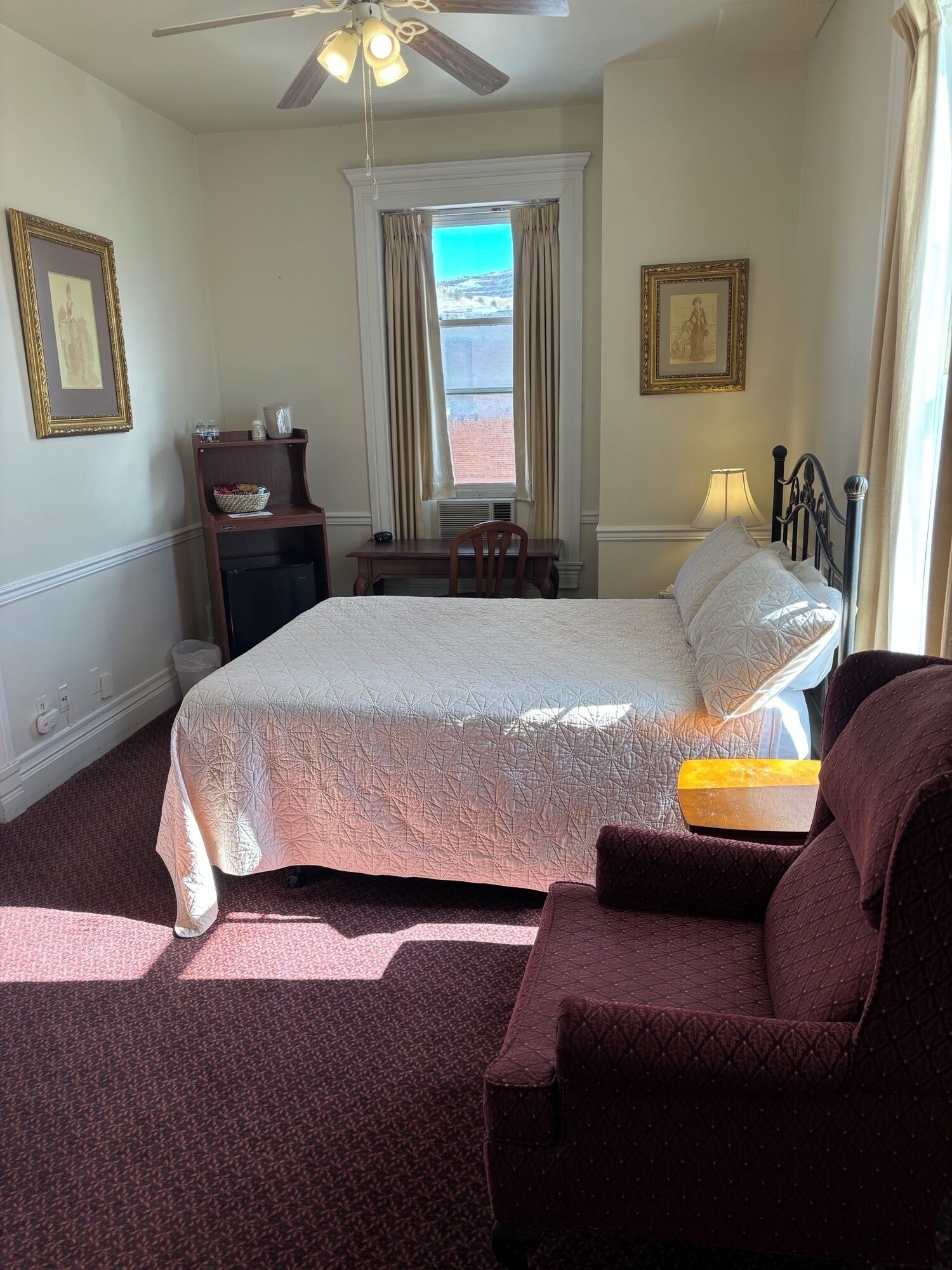 Ein Hotelzimmer mit Bett, Schreibtisch, Stuhl, Deckenventilator und einem Fenster mit Vorhängen.