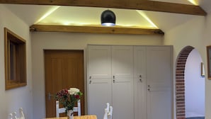 Un comedor con una mesa de madera, sillas blancas y un jarrón de flores. Hay armarios blancos y una viga de techo de madera.