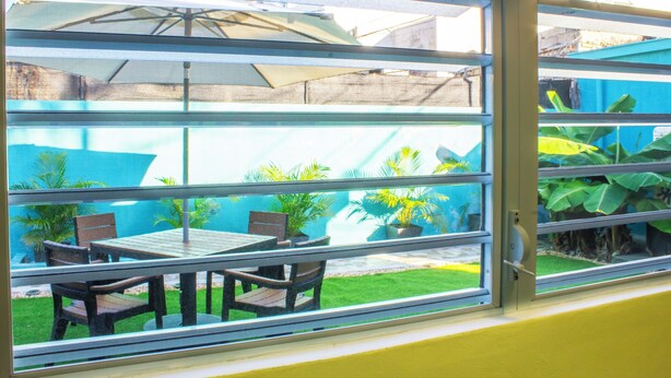 A window with a view of an outdoor patio area featuring a table and chairs, a potted plant, and a blue building in the background.