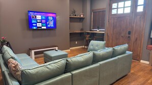 A modern living room with a sectional sofa, a flat-screen TV mounted on the wall, a wooden dining table, and a kitchen area in the background. - NEW! Modern Private Entrance, 2BD Apt. Minutes to NYC (Jersey City)