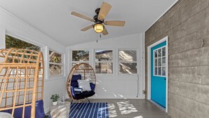 Une véranda couverte avec un ventilateur de plafond, une chaise en osier, une balançoire rembourrée et un tapis à rayures bleues.