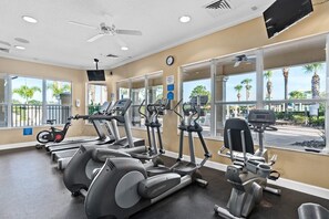 Une salle de sport équipée de tapis de course, de vélos elliptiques et d’un vélo d’appartement, de grandes fenêtres offrant une vue sur les palmiers et d’un ventilateur de plafond.