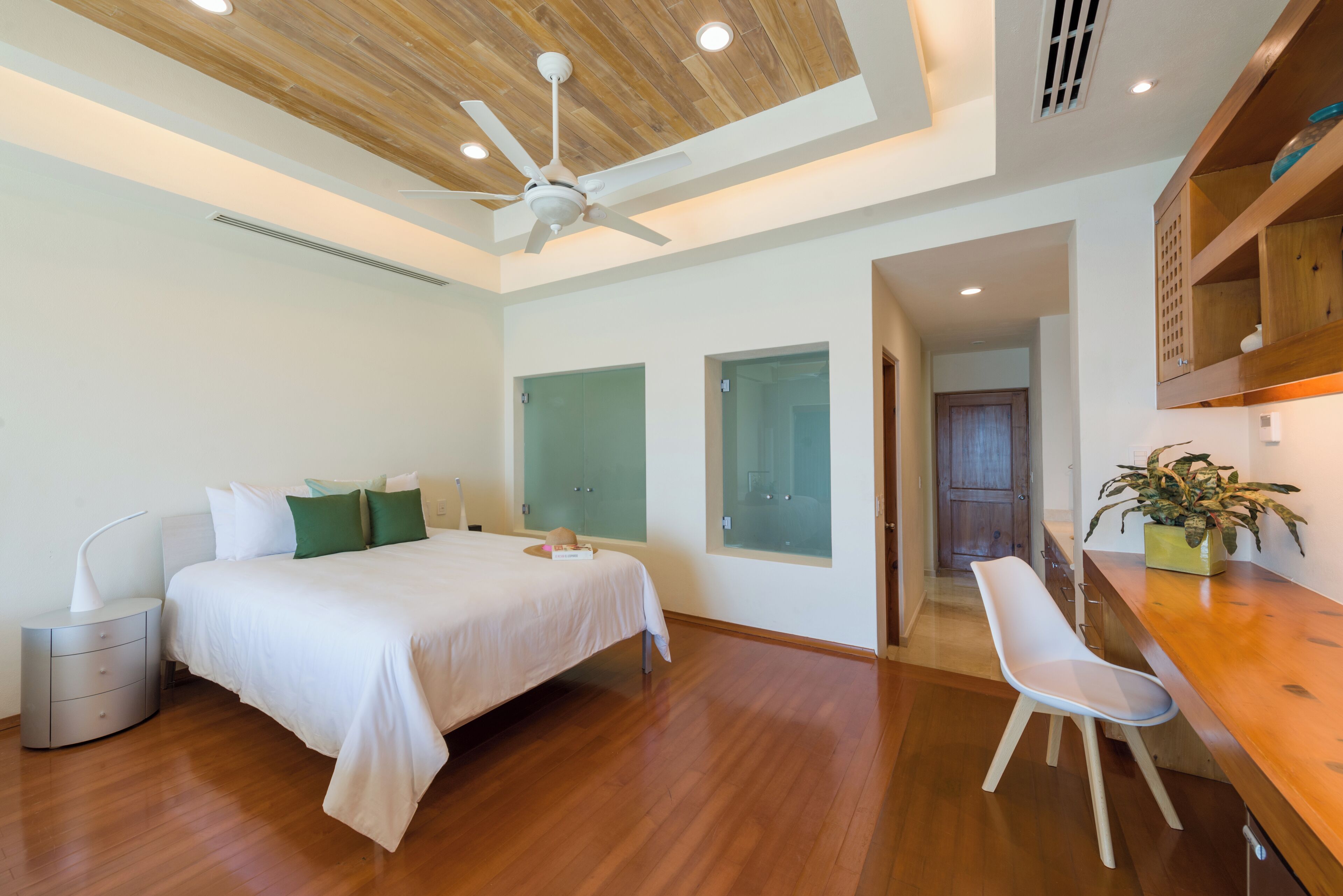 A bedroom with a bed, bedside table, nightstand, ceiling fan, and a wooden desk with a chair.