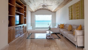 A spacious living room with a large window, a ceiling fan, a sofa with cushions, and a wooden bookshelf. - Villa Punta Sam Cancun Beachfront Luxury (Cancun)