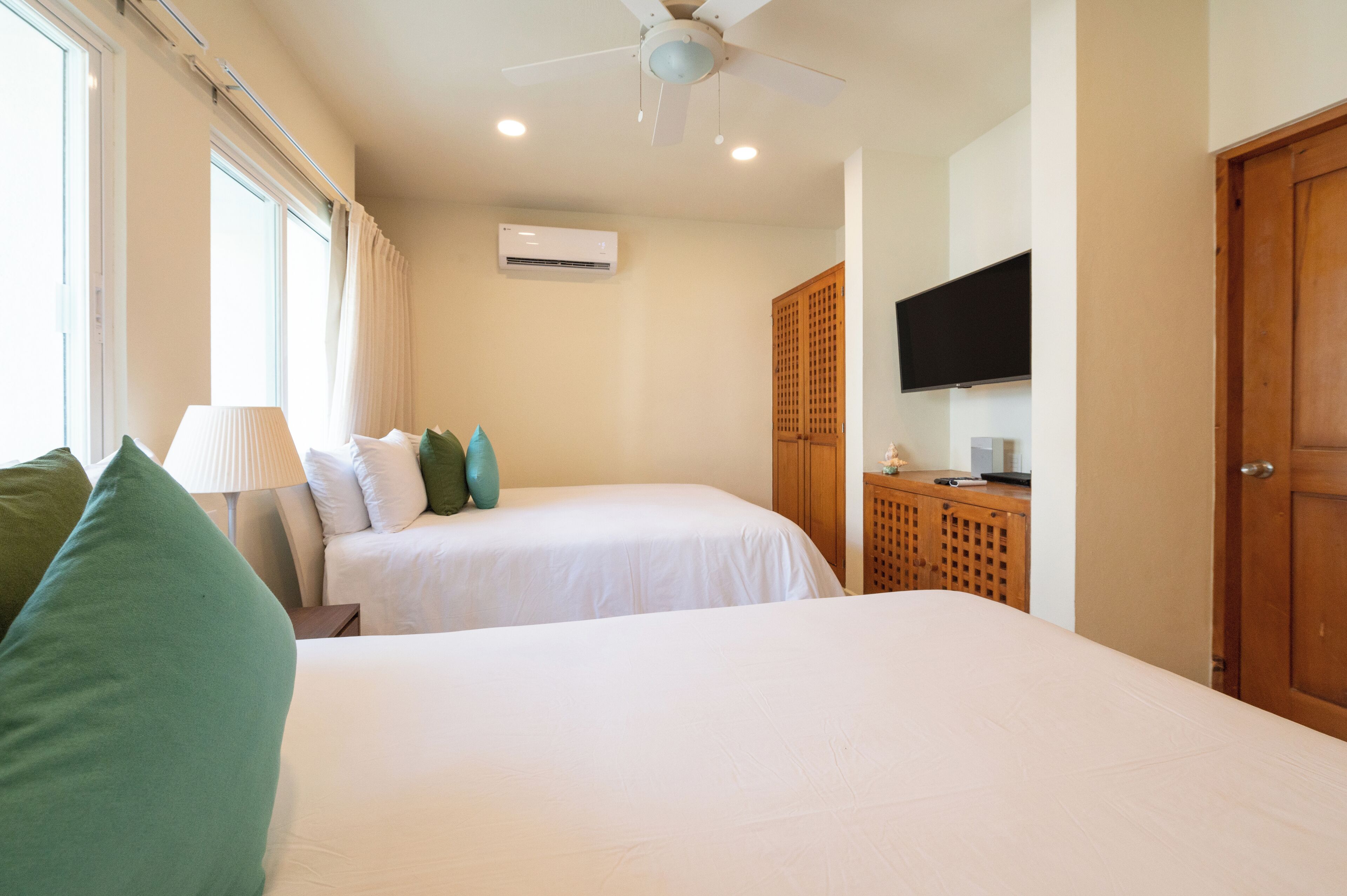 Habitación de hotel con dos camas, ventilador de techo, unidad de aire acondicionado, televisor sobre un mueble de madera y una ventana con cortinas.