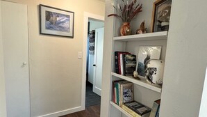 A hallway with a bookshelf containing books and decorative items, a framed picture on the wall, and a door leading to another room. - Newly Remodeled Charming Cozy 3-bedroom house in Anchorage with WiFi (Anchorage)
