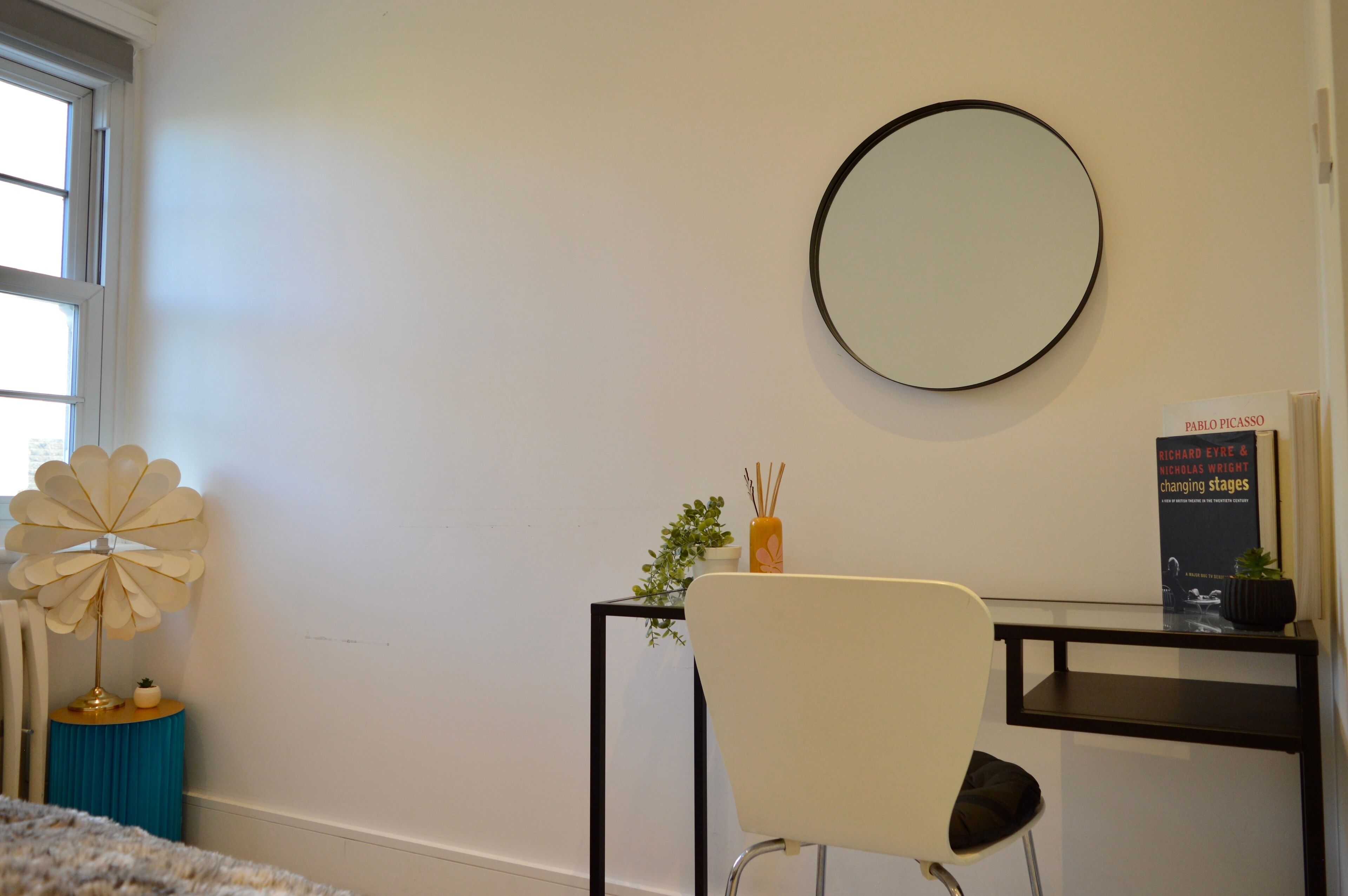 A room with a round mirror on the wall, a white chair, a small table with a plant, and a book titled 'Changing Stages' by Richard Eyre & Nicholas Wright.
