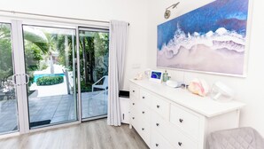 Una habitación con puerta corrediza de vidrio que da a un patio, un aparador blanco con cajones, un cuadro enmarco y una pintura de una ola.