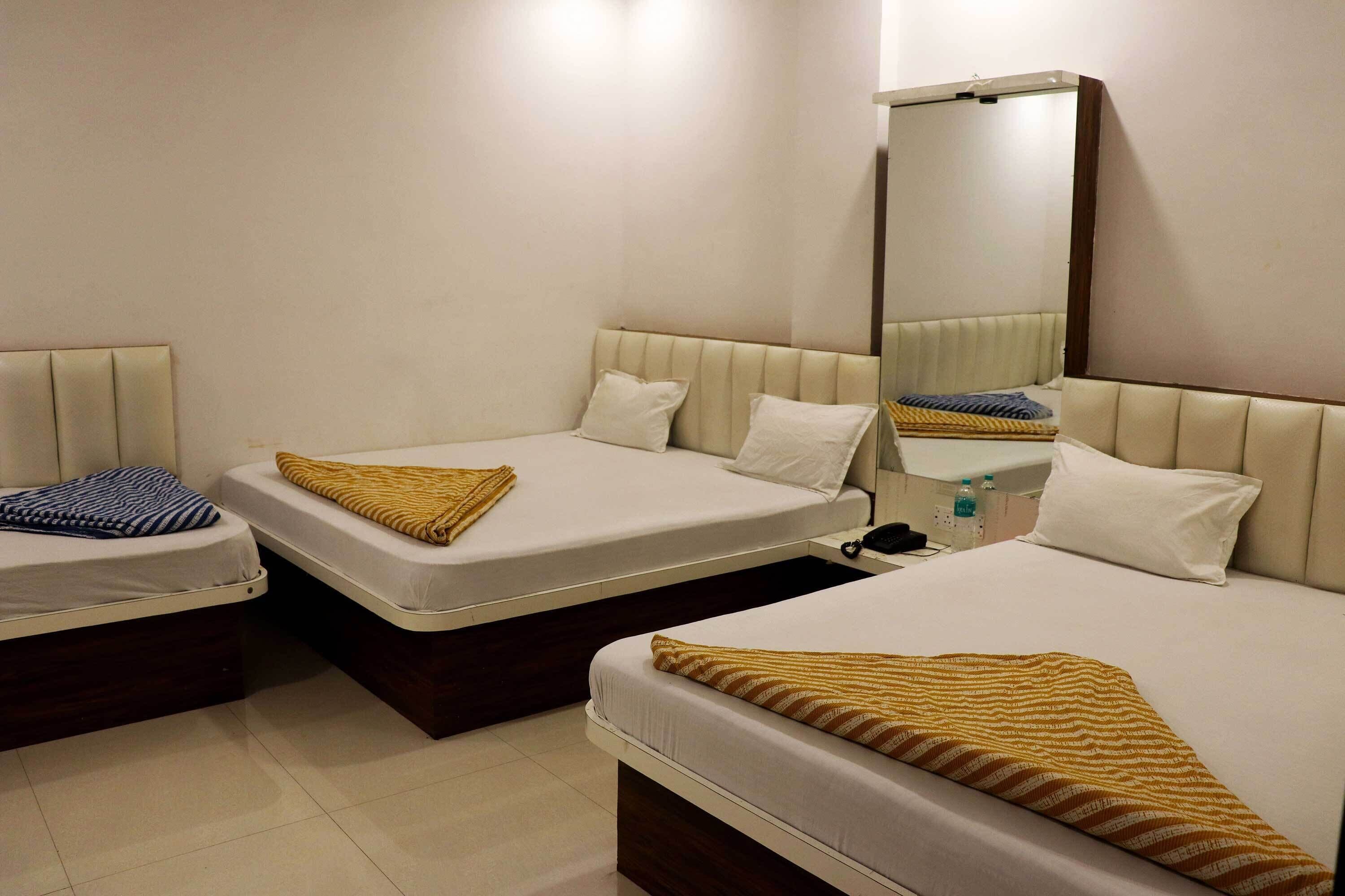 Une chambre d’hôtel avec trois lits, chacun recouvert d’une couverture à motifs différents. Il y a un miroir au mur et une petite table sur laquelle se trouvent un téléphone et une bouteille.