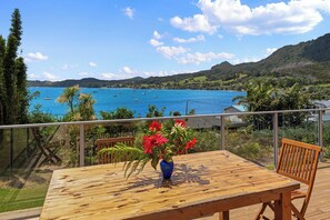 Une terrasse en bois avec une table et des chaises, donnant sur une baie côtière où l’on peut voir des bateaux et une végétation luxuriante.