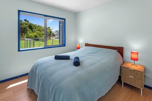 Une chambre avec une grande fenêtre, un lit recouvert d’une couverture bleue, deux lampes de chevet et une table de chevet en bois.