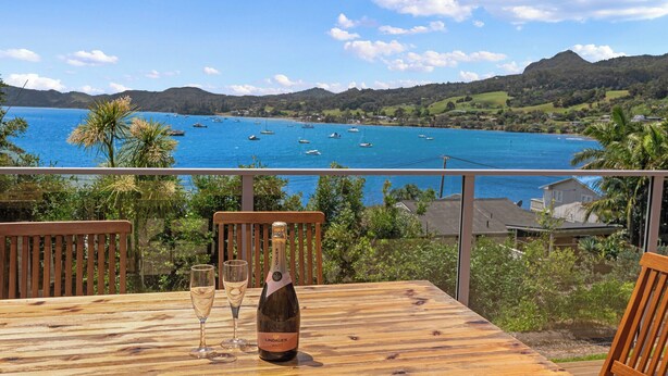 Eine Holzterrasse mit einer Flasche Lindauer Fruchtwein und zwei Gläsern, mit Blick auf eine Küstenbucht, in der Boote und Hügel zu sehen sind.