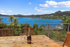 Eine Holzterrasse mit einer Flasche Lindauer Fruchtwein und zwei Gläsern, mit Blick auf eine Küstenbucht, in der Boote und Hügel zu sehen sind.