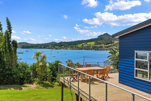 Une terrasse dotée de mobilier d’extérieur, offrant une vue sur une baie où sont amarrés des bateaux, le tout entouré de verdure et de collines.