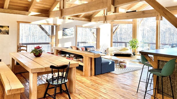 A spacious room with wooden beams, large windows, a wooden dining table, and a sofa. - Arete Hostel (Summersville)