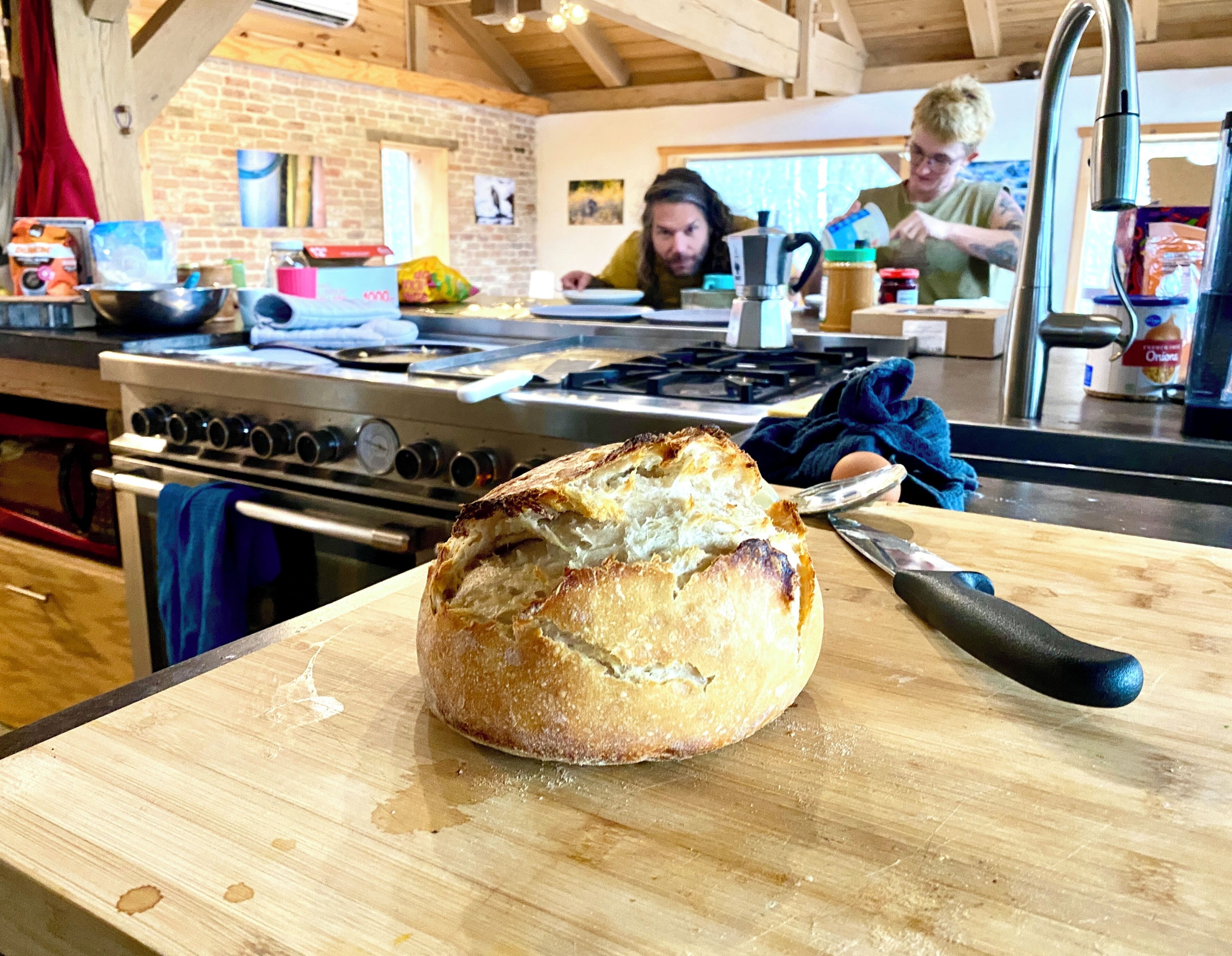 Uma barra de pão sobre uma tábua de corte de madeira em uma cozinha, com um fogão e diversos utensílios domésticos ao fundo.