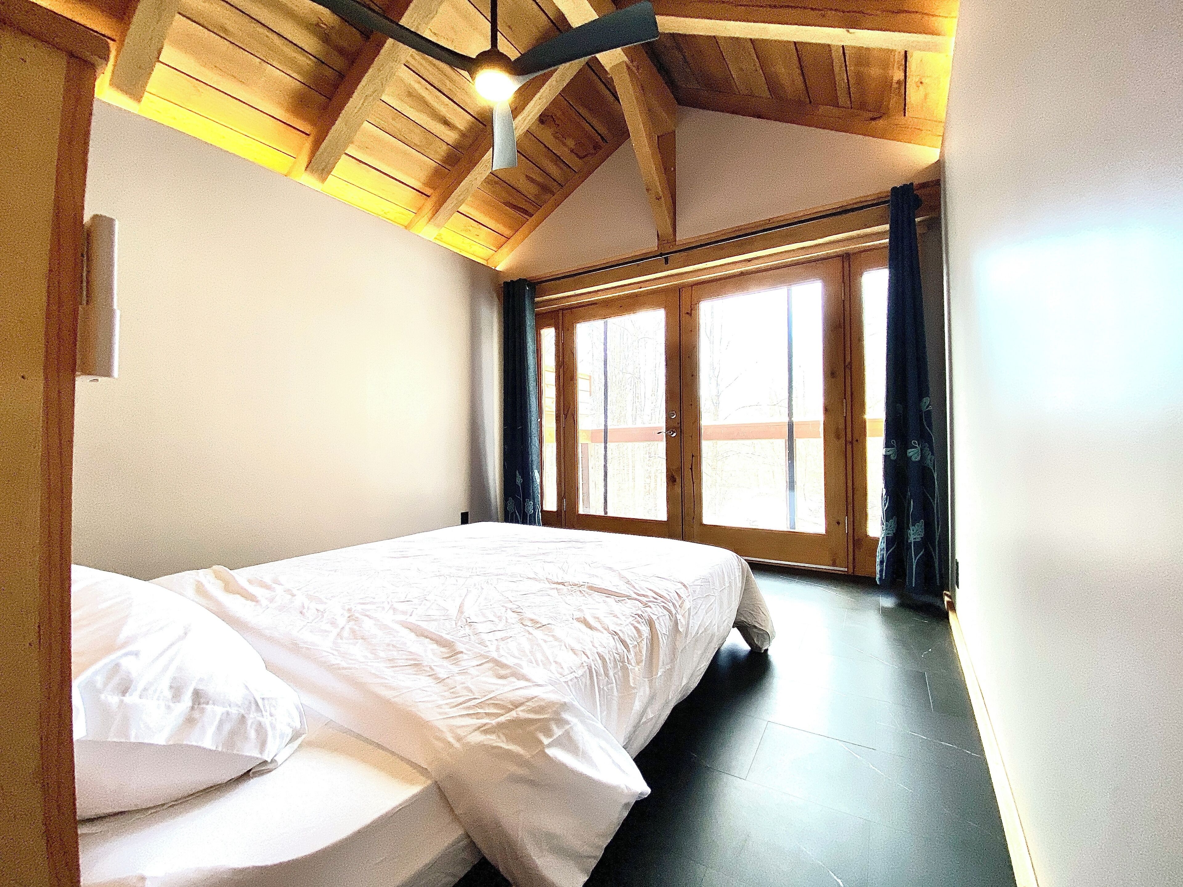 Un dormitorio con cama, vigas de madera en el techo y un ventanal con cortinas.