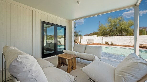 Zona salotto esterna coperta con divani bianchi imbottiti e un tavolo in legno, con vista su una piscina e un paesaggio desertico.
