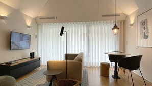 Un salon moderne avec un canapé, une petite table ronde, un téléviseur fixé au mur et une grande fenêtre ornée de rideaux légers.