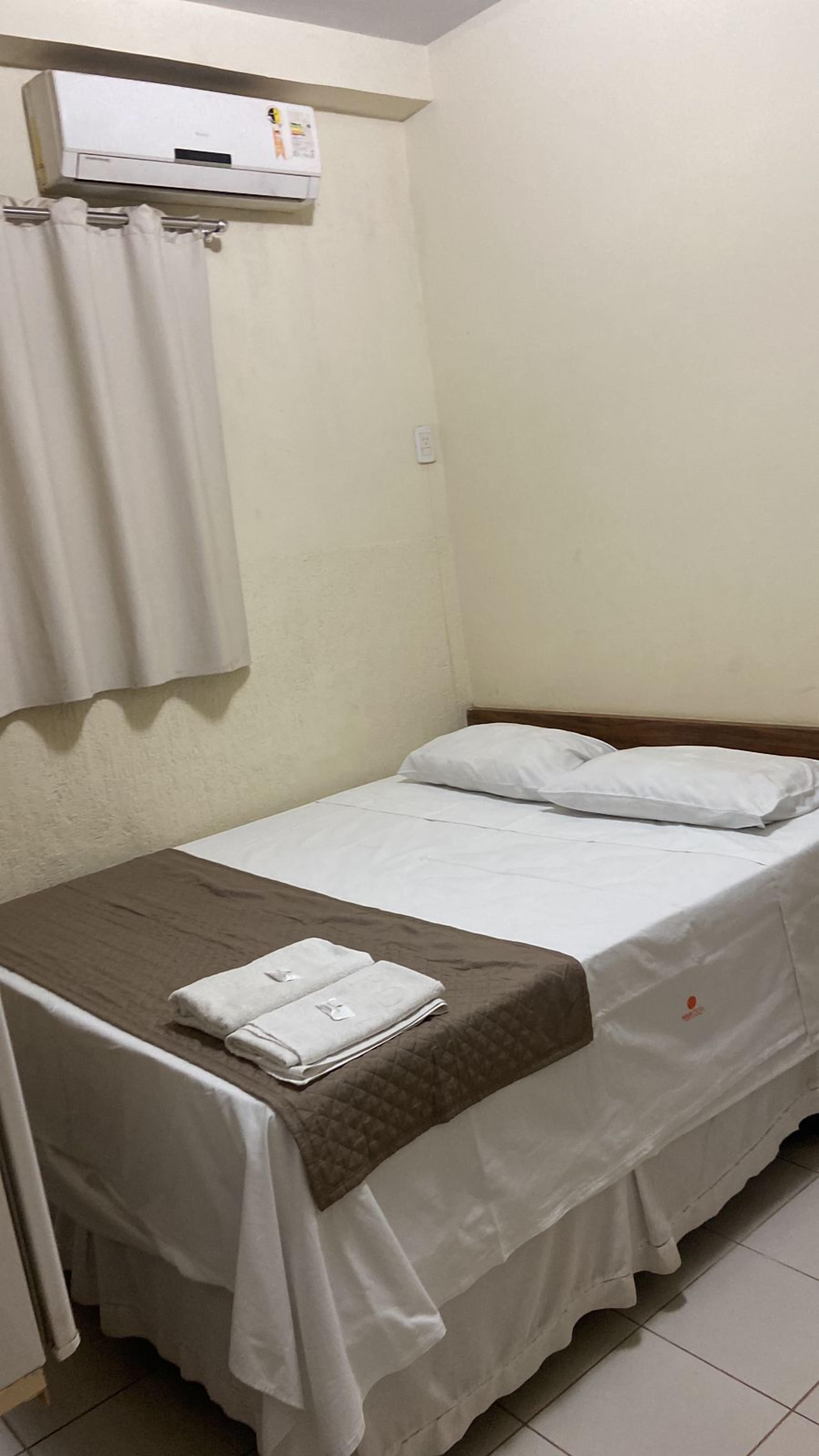 Une petite chambre avec un lit simple, une tête de lit en bois, un couvre-lit blanc et une couverture matelassée marron. Il y a un rideau blanc sur le côté gauche et un climatiseur fixé au mur au-dessus du lit.