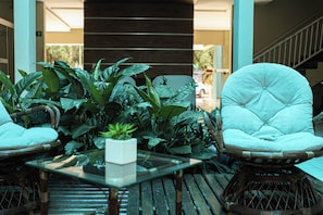 Una silla de mimbre con asiento y respaldo acolchados, una mesita de centro de vidrio con una planta en maceta, y suelo de madera.