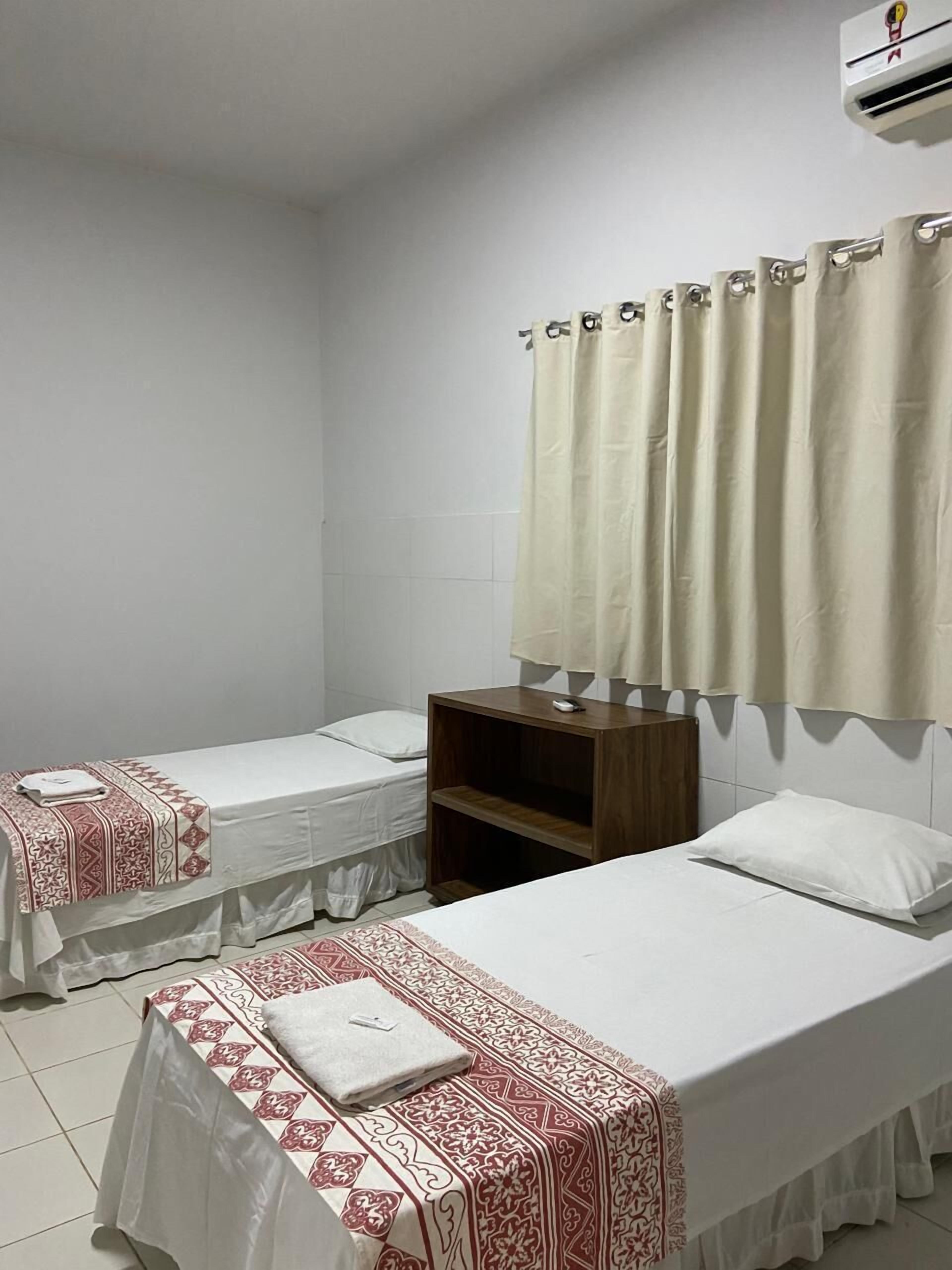 A hotel room with two single beds, a wooden nightstand, a white air conditioning unit, and a window with beige curtains.