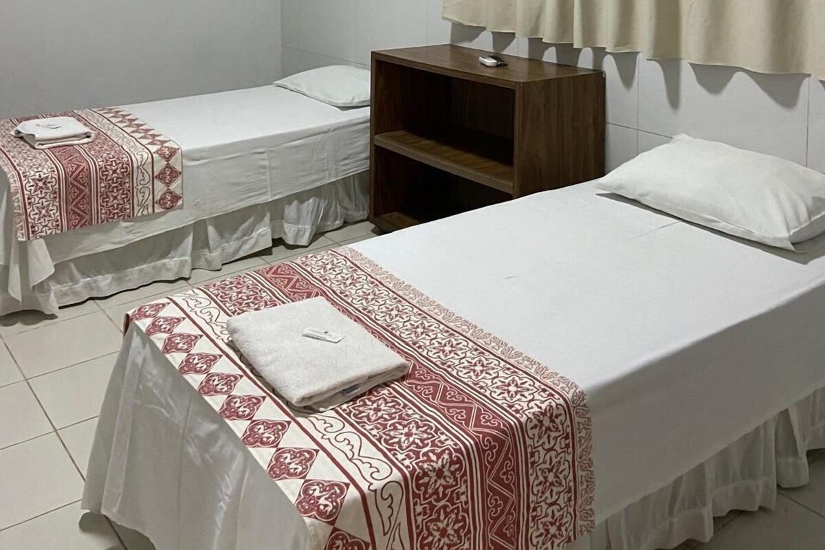 A hotel room with two single beds, a wooden nightstand, a white air conditioning unit, and a window with beige curtains.