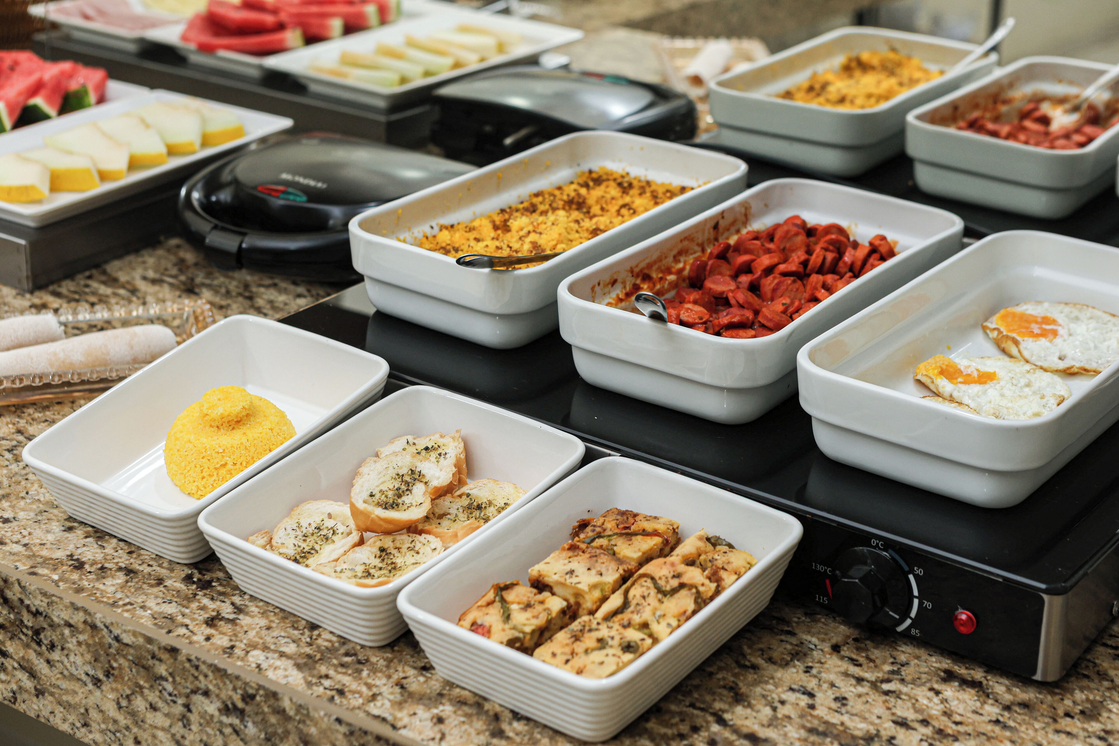 A buffet with various food items in white rectangular trays, including bread, eggs, and meats.